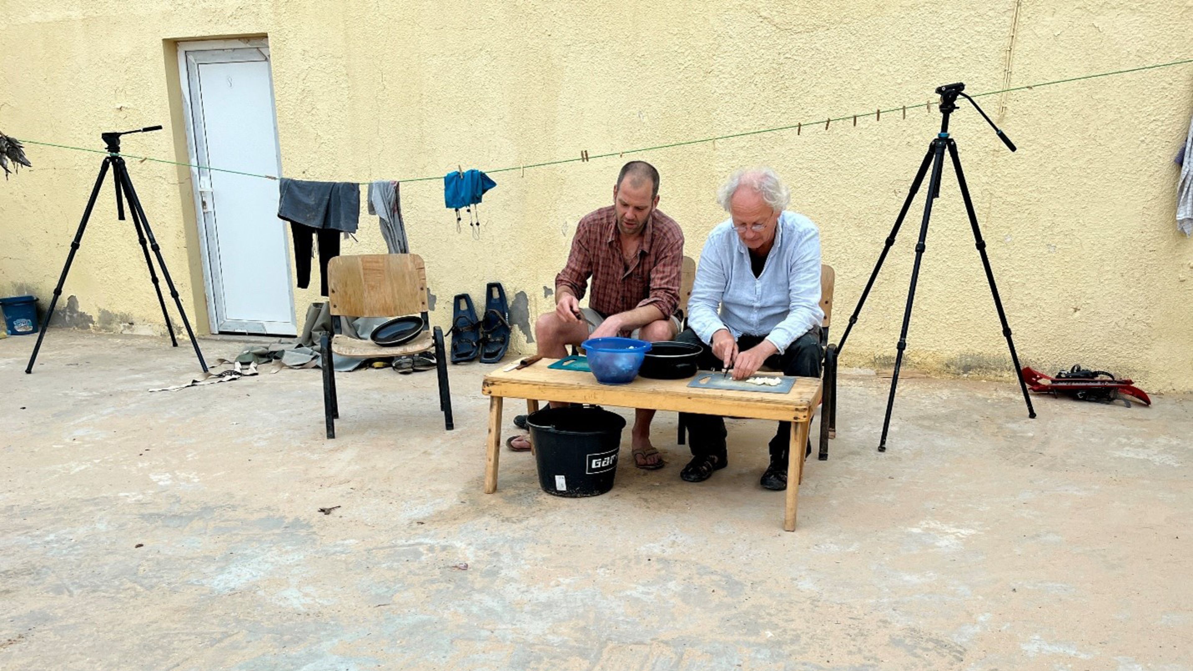 Photo of two persons (Roeland Bom and Theunis Piersma) sitting behind a table in front of a light yellow wall, discussing science while preparing dinner in Banc d’Arguin, Mauritania